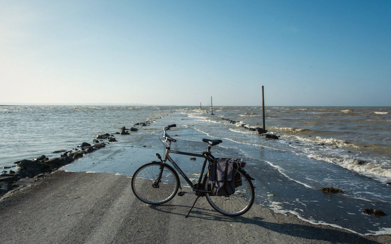 A parked bicycle with panniers stands on a narrow causeway flanked by waves on both sides