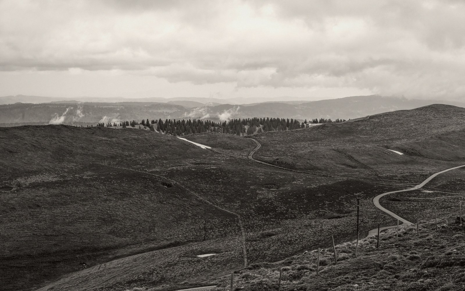 A winding road crosses a hilly plateau beneath cloudy skies, with distant tree lines and lingering snow patches