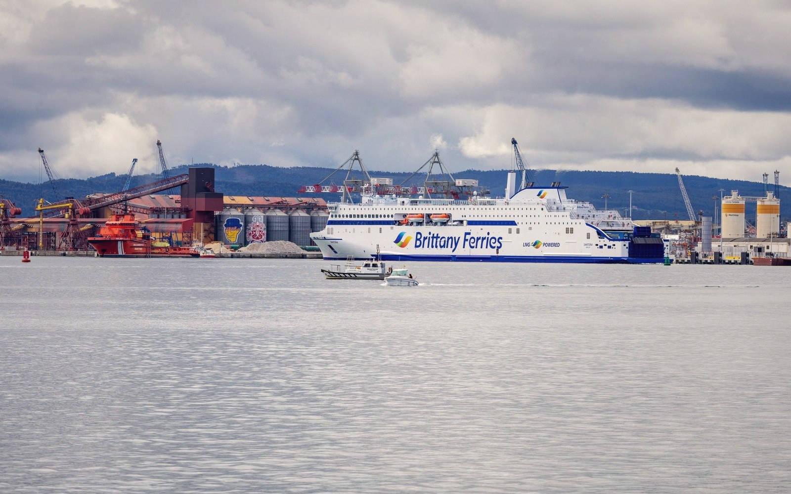 A Brittany Ferries ship docked at a port beside industrial cranes and silos