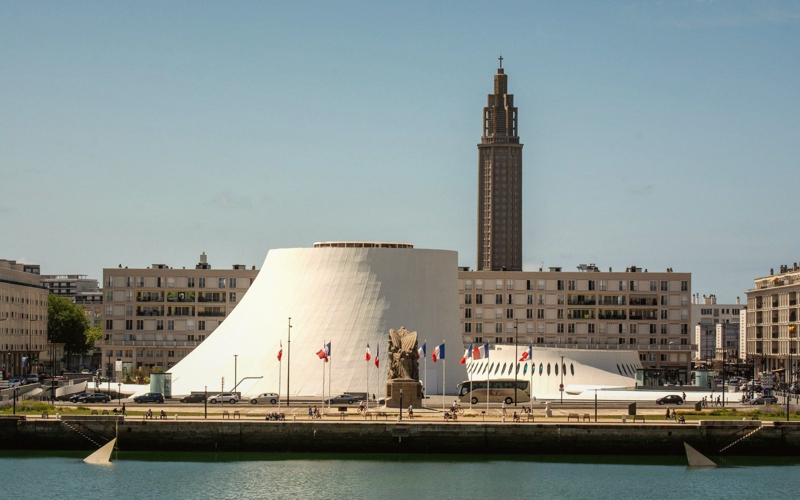A cylindrical white cultural building stands beside a tall church tower along the waterfront