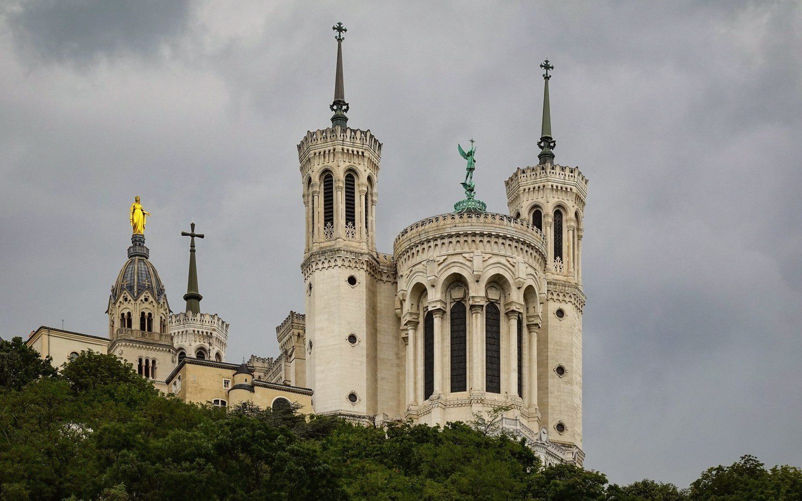 Basilica towers with spires and a golden statue rise above a hillside of trees