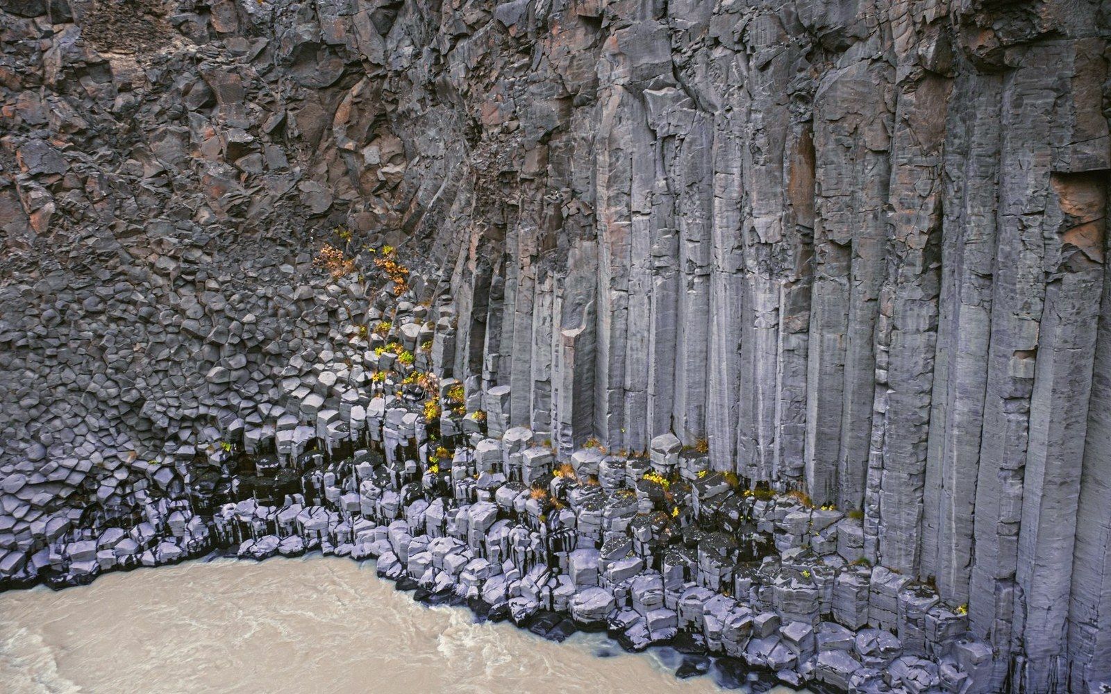 Basalt columns rise above a muddy river, with small shrubs growing between the rock formations