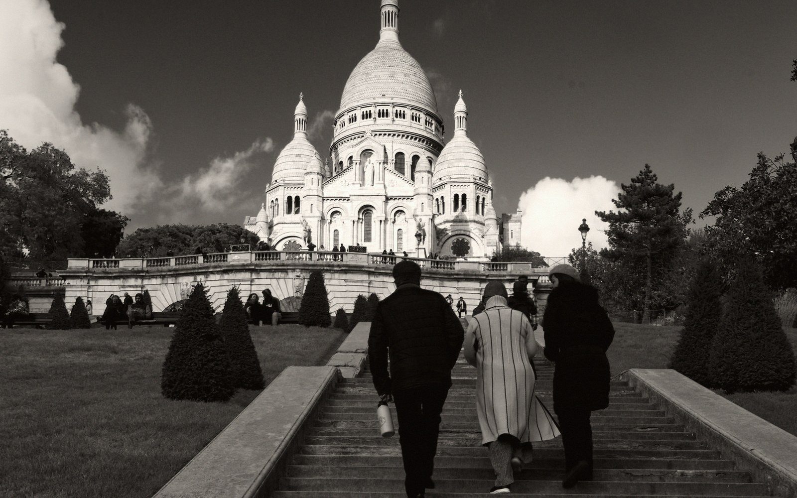 The broad stone steps below Sacré-Cœur in morning light, adults sitting and walking, Paris rooftops below